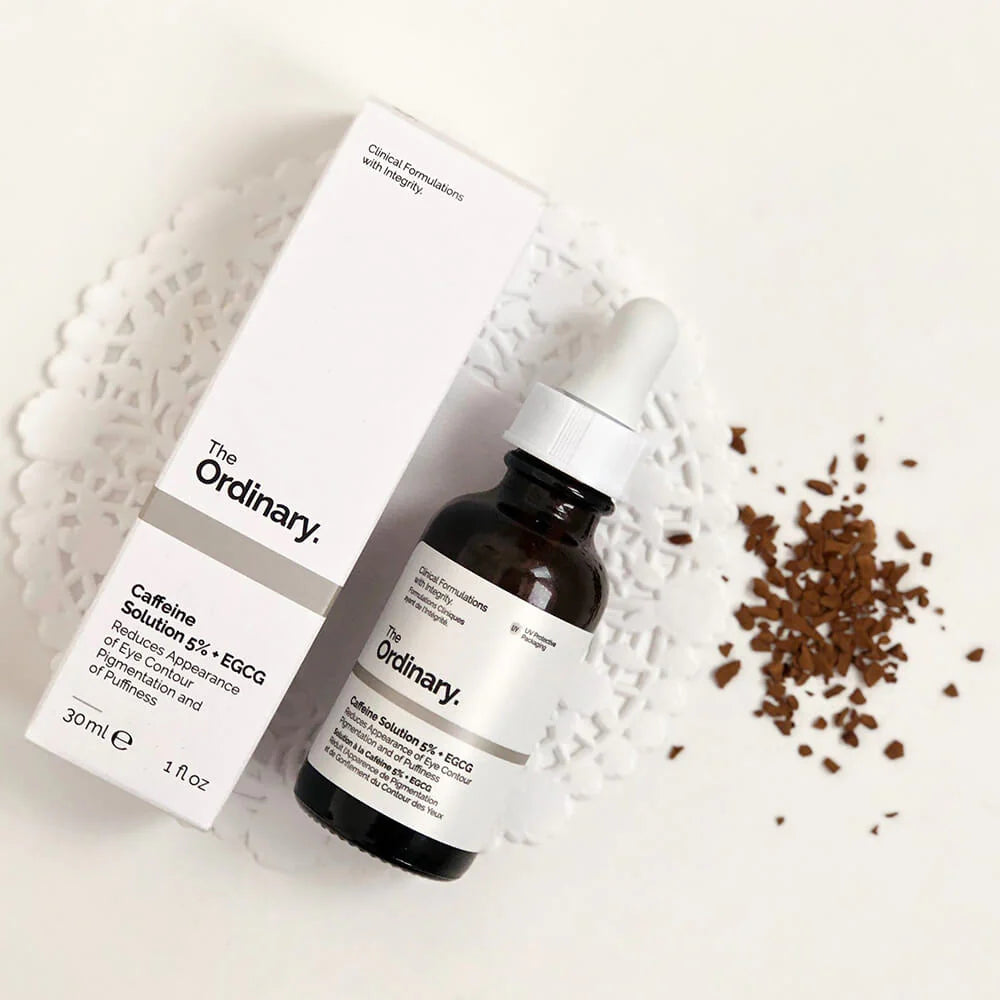 The Ordinary skincare serum bottle and packaging on a textured white surface with scattered coffee beans.

